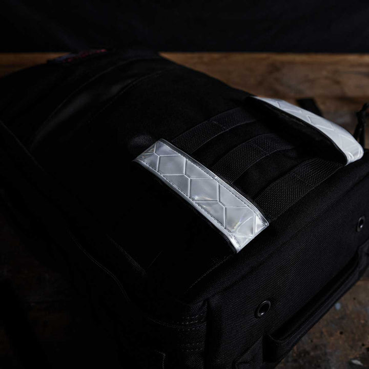 A GORUCK black backpack with quilted silver straps, featuring their signature Reflective Ruck Bands for enhanced nighttime visibility, rests elegantly on a wooden surface.