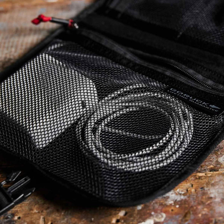 A Wire Dopp by GORUCK rests on a wooden surface, its open mesh pouch neatly holding a white power adapter and coiled cable for organized carry. This pouch is part of a durable black CORDURA bag, featuring a convenient zipper with a red pull tab—an ideal companion for your travel essentials.