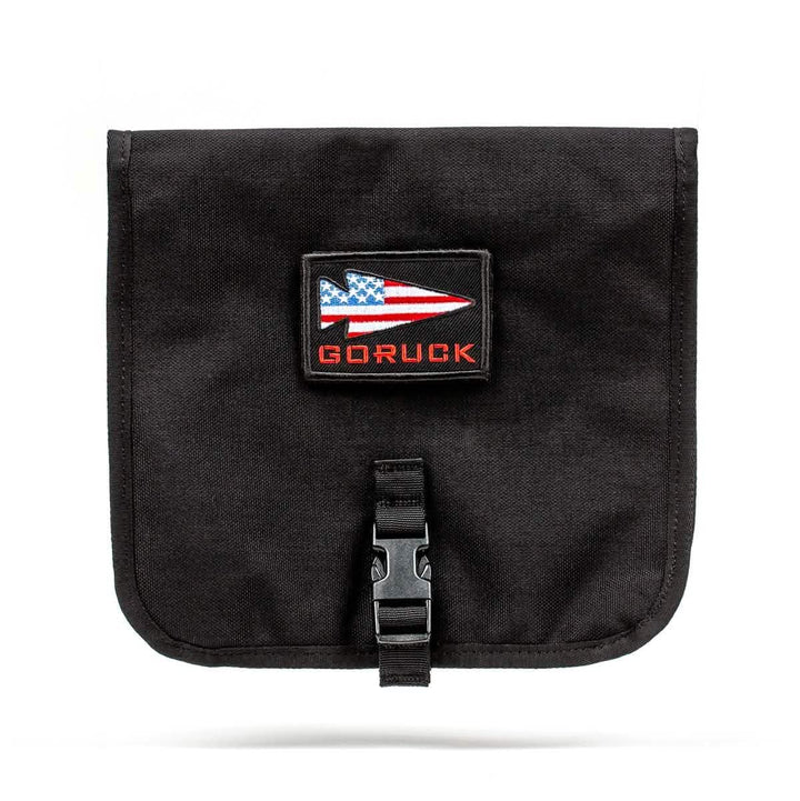 Introducing the GORUCK Wire Dopp: a black pouch designed for organized carry, featuring a flap adorned with a rectangular patch of an American flag and the brand name "GORUCK" in bold red. The pouch is secured with a black buckle closure beneath the flap, enhancing its rugged durability. Set against a white background, this product perfectly combines style and functionality.