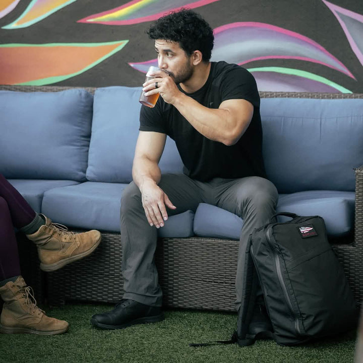 A man sits on a couch, sipping a drink with his waterproof GORUCK GR1 USA - X-PAC backpack featuring AquaGuard zippers beside him, set against a vibrant mural backdrop.