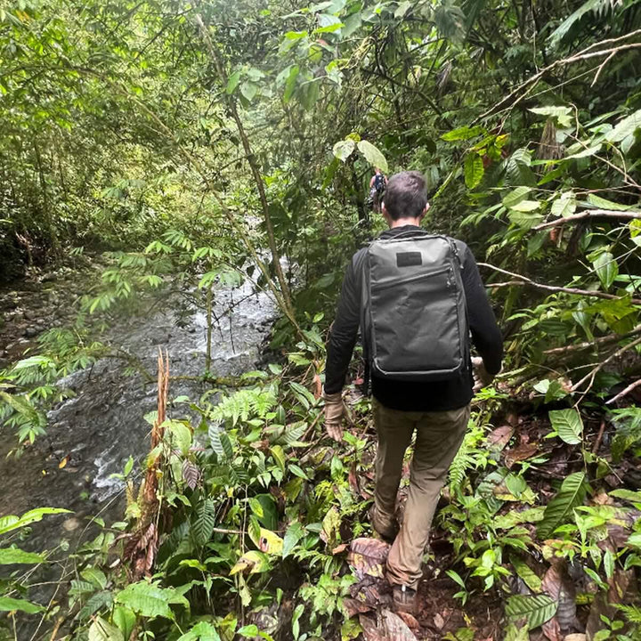 A person carrying the GORUCK GR1 USA - X-PAC backpack confidently strolls through a lush jungle by a flowing stream, trusting the AquaGuard zippers to ensure their gear stays waterproof and dry.